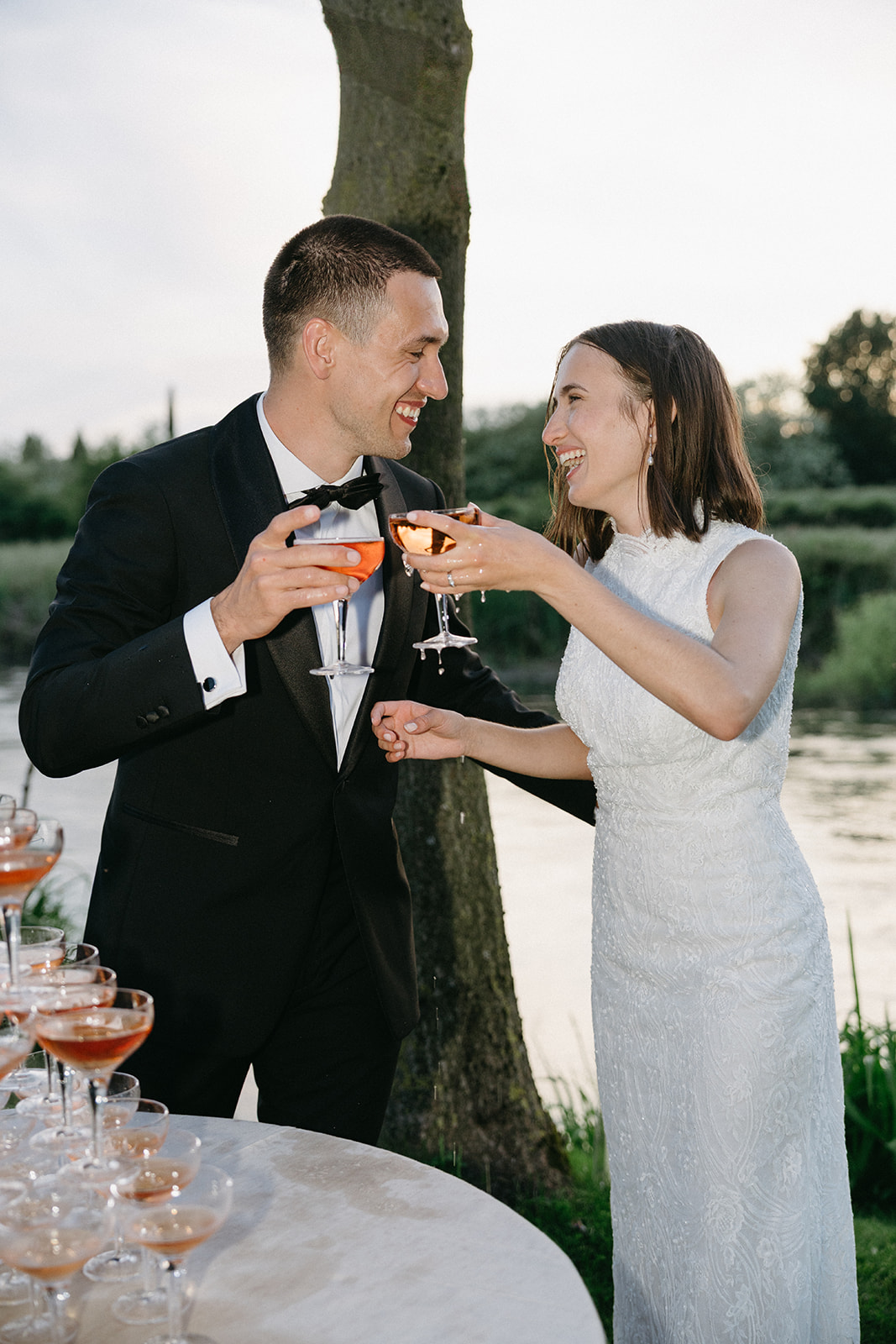 Bride and groom laughing while toasting with rosé by the water during their wedding in Tuscany